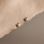 Two Baby Venus Flatback Studs in Silver, featuring delicate pearl earrings, are displayed on a soft beige fabric surface.