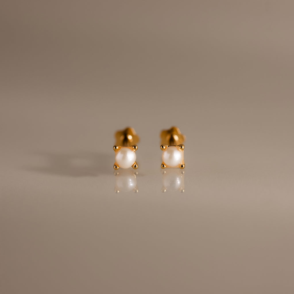 A pair of Baby Venus Flatback Studs in 18K Gold, featuring delicate pearls and floral backing, displayed on a smooth, reflective surface.