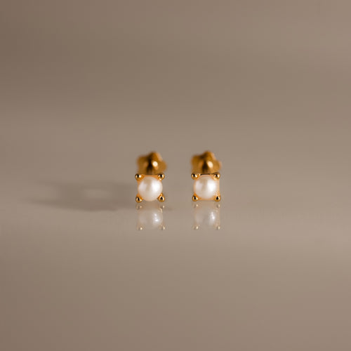 A pair of Baby Venus Flatback Studs in 18K Gold, featuring delicate pearls and floral backing, displayed on a smooth, reflective surface.