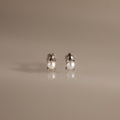 The Baby Venus Flatback Studs in Silver are delicate pearl earrings with silver prongs and a floral backing, displayed on a reflective beige surface.