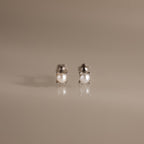 The Baby Venus Flatback Studs in Silver are delicate pearl earrings with silver prongs and a floral backing, displayed on a reflective beige surface.