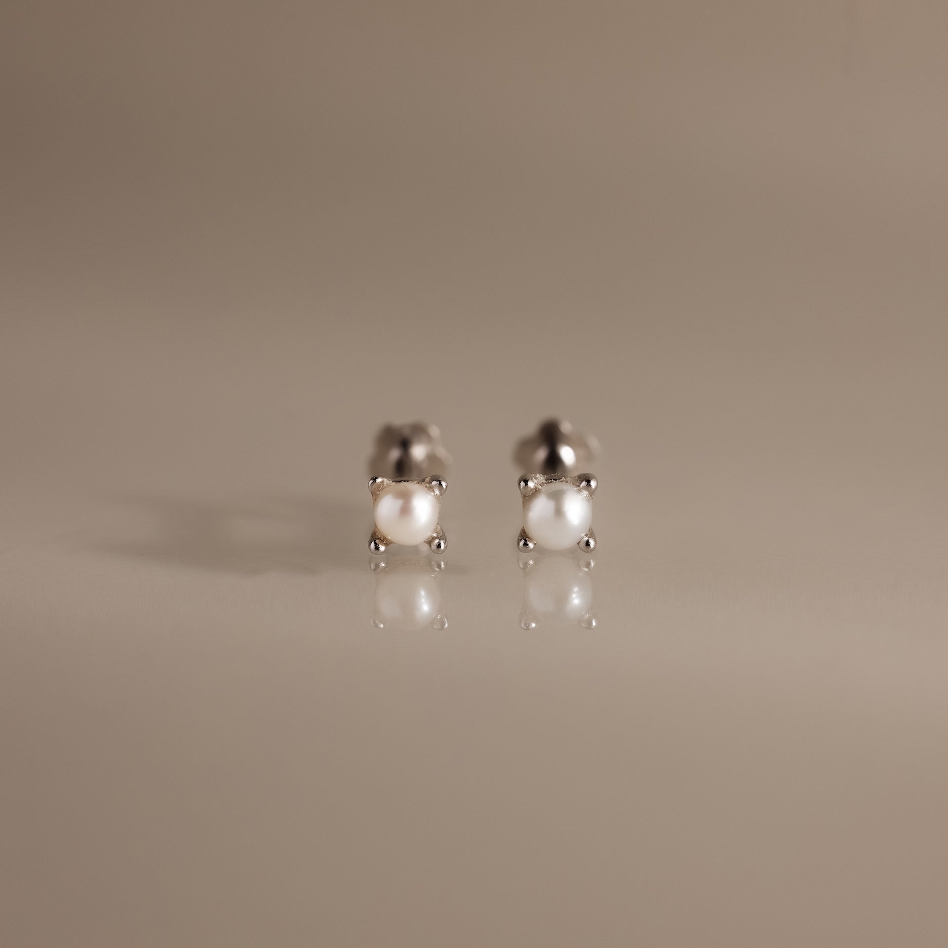 The Baby Venus Flatback Studs in Silver are delicate pearl earrings with silver prongs and a floral backing, displayed on a reflective beige surface.
