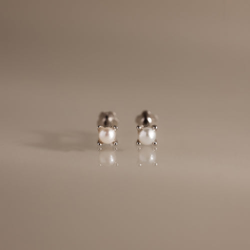 The Baby Venus Flatback Studs in Silver are delicate pearl earrings with silver prongs and a floral backing, displayed on a reflective beige surface.