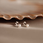 The Baby Venus Flatback Studs in Silver, two petite pearl earrings with silver backing, are displayed beside a seashell on a beige surface.