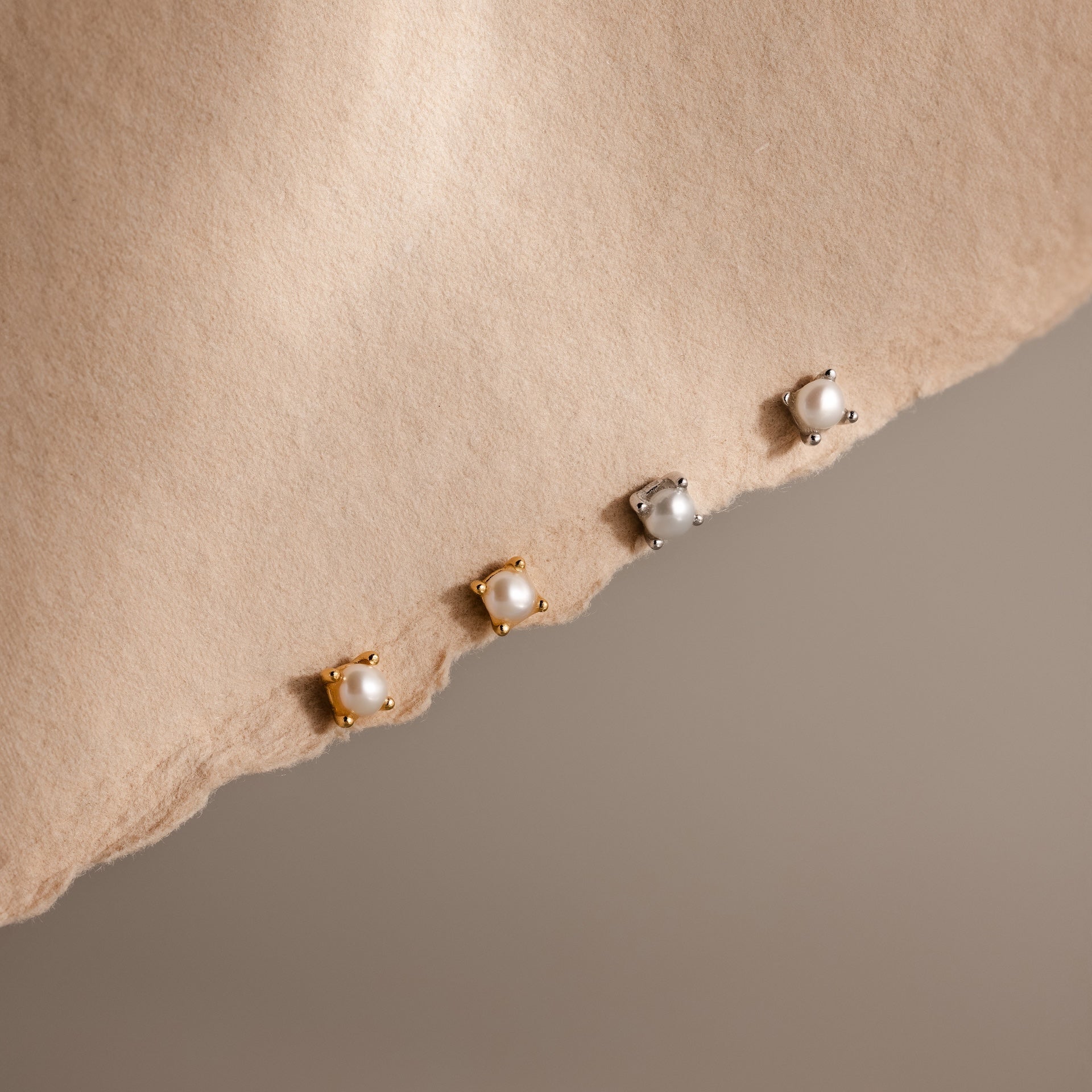 Three pairs of Baby Venus Flatback Studs—two gold, one silver—rest on a torn beige paper background, creating an elegant jewelry set with timeless charm for any look.
