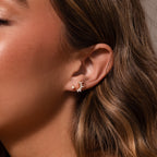 Close-up of a womans ear with elegant jewelry—two gold earrings, including the Baby Venus Flatback Studs and a leaf-shaped cluster with floral backing.