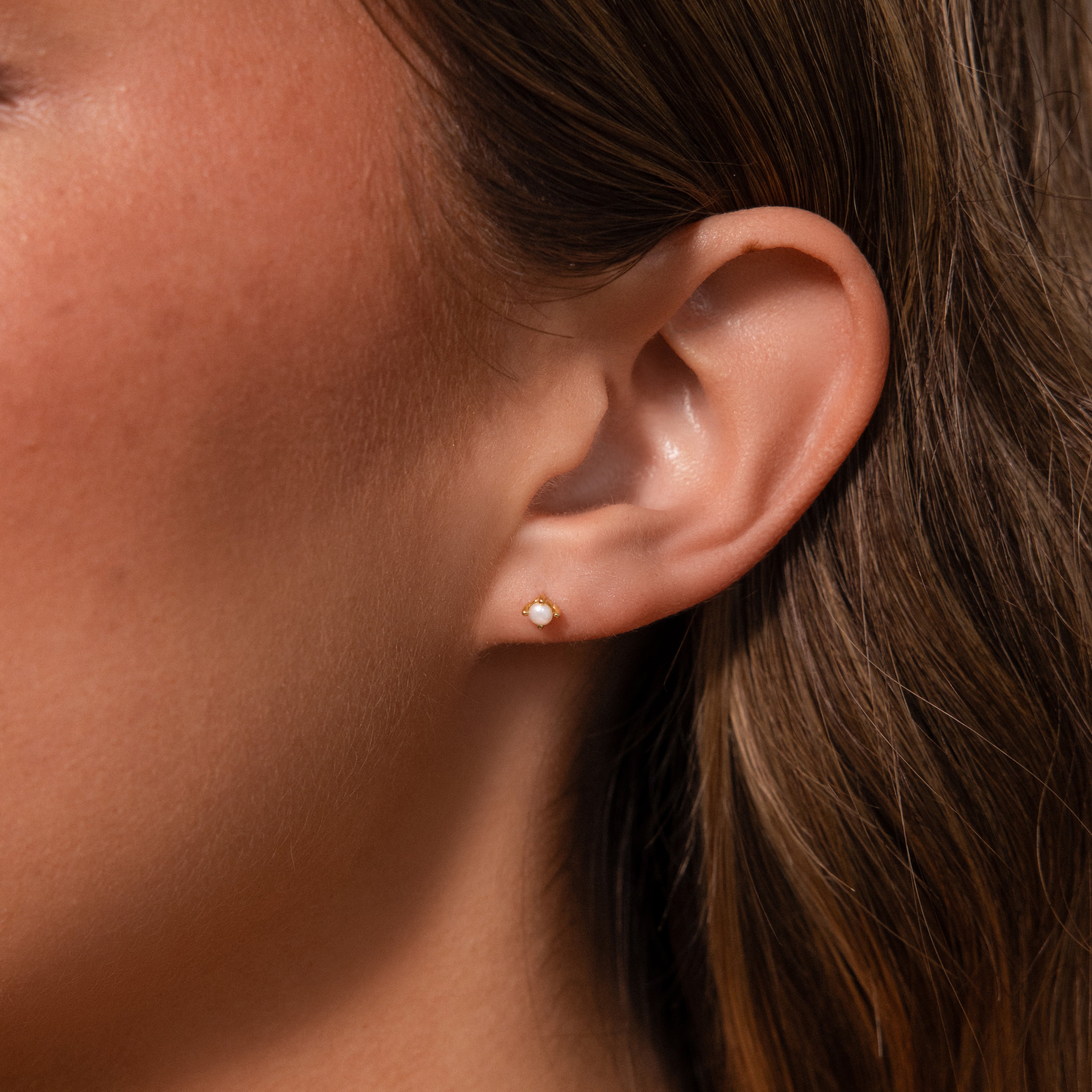 Close-up of a womans ear wearing Baby Venus Flatback Studs in 18K Gold with a floral backing, her light brown hair softly framing the delicate pearls.