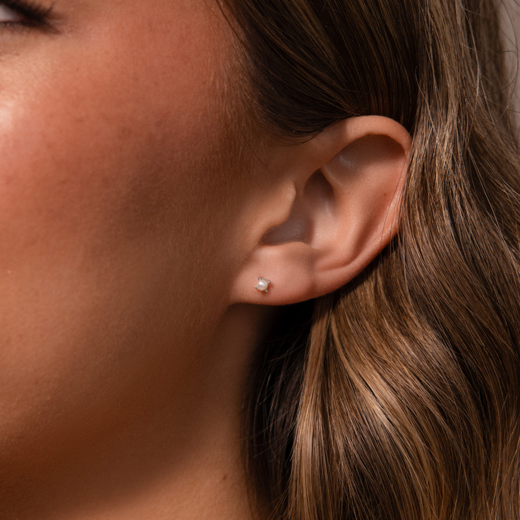 Close-up of a womans ear with wavy brown hair, showcasing Baby Venus Flatback Studs in Silver and highlighting the delicate pearl earrings with subtle floral backing.