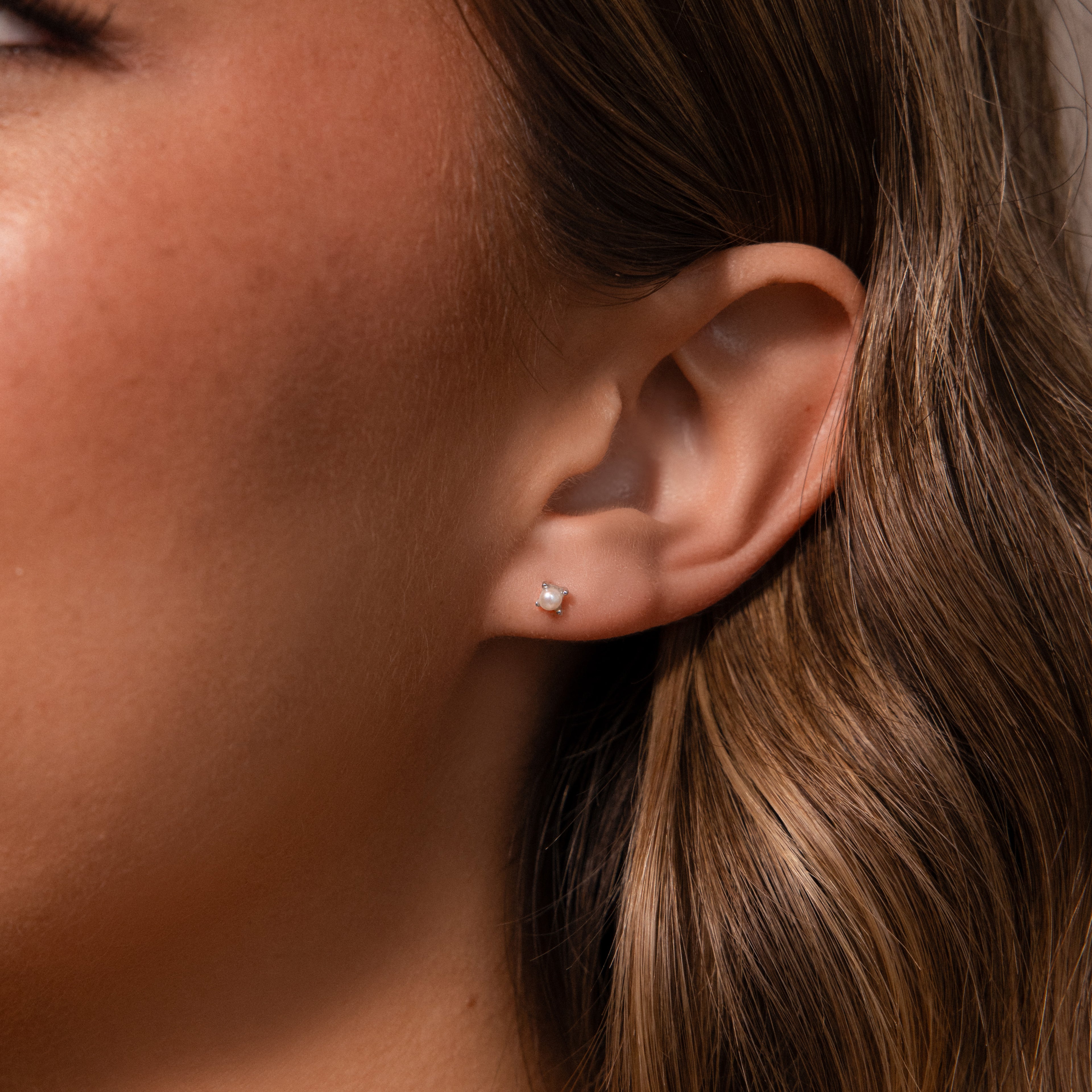 Close-up of a womans ear with wavy brown hair, showcasing Baby Venus Flatback Studs in Silver and highlighting the delicate pearl earrings with subtle floral backing.