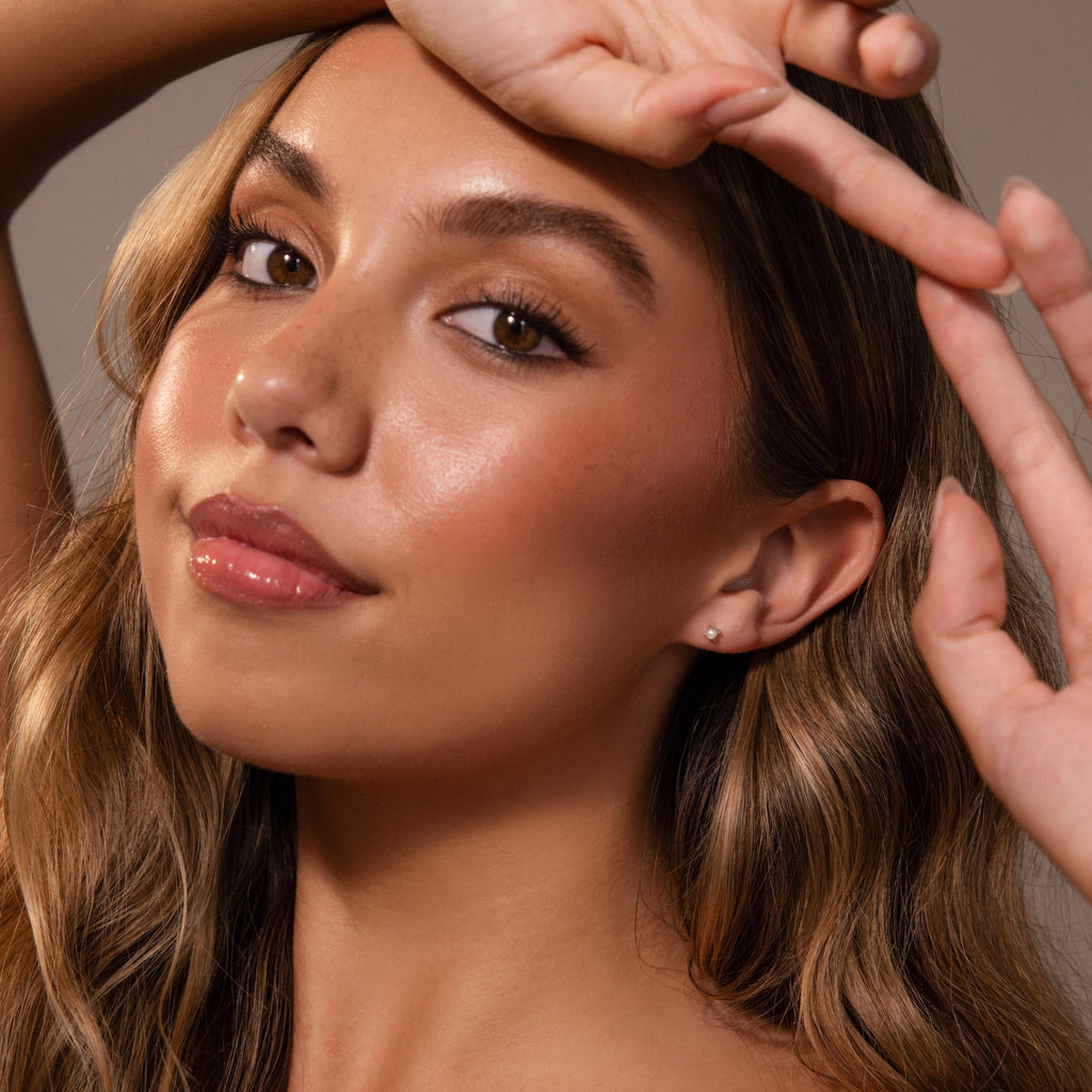 A woman with long wavy hair poses with one arm raised, showcasing glowing skin, natural makeup, and Baby Venus Flatback Studs in Silver.