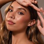A woman with long wavy hair poses with one arm raised, showcasing glowing skin, natural makeup, and Baby Venus Flatback Studs in Silver.