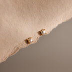 Baby Venus Flatback Studs in 18K Gold, featuring delicate pearls and floral backing, are displayed on a soft beige fabric background.