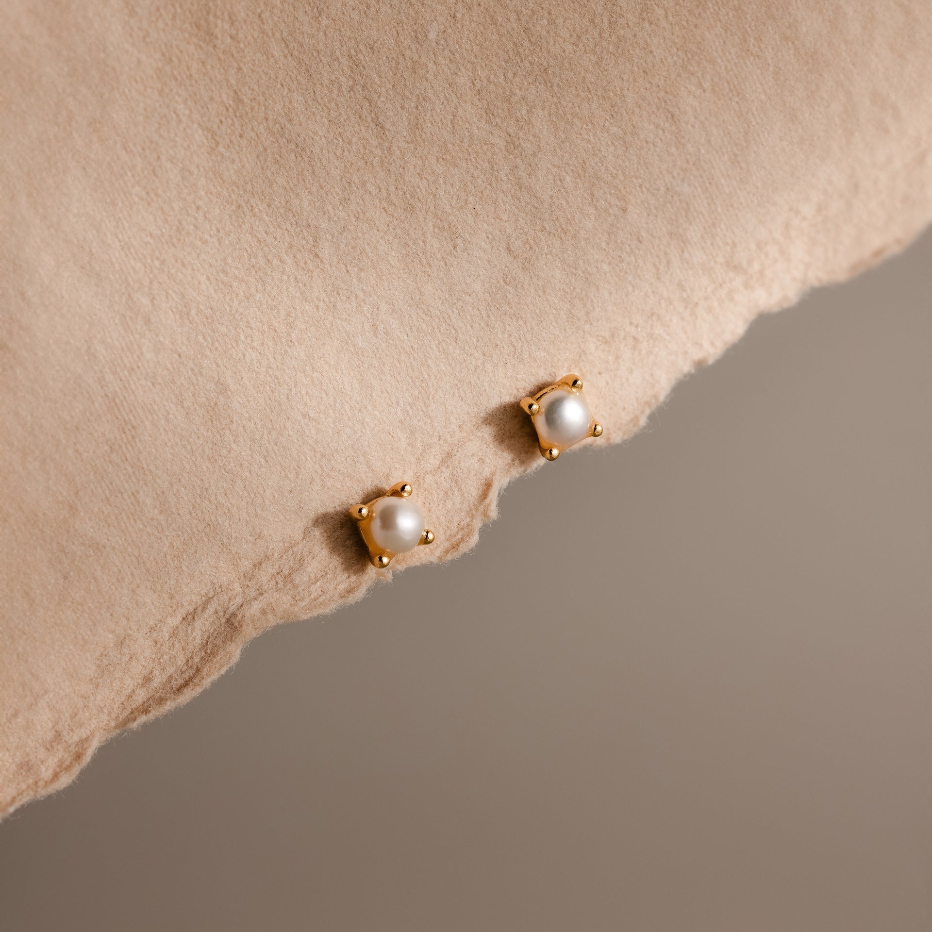 Baby Venus Flatback Studs in 18K Gold, featuring delicate pearls and floral backing, are displayed on a soft beige fabric background.