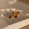 Elegant Garnet Flatback Studs with red gemstones, these gold earrings add a dainty touch and look stunning against a textured, light-colored surface.