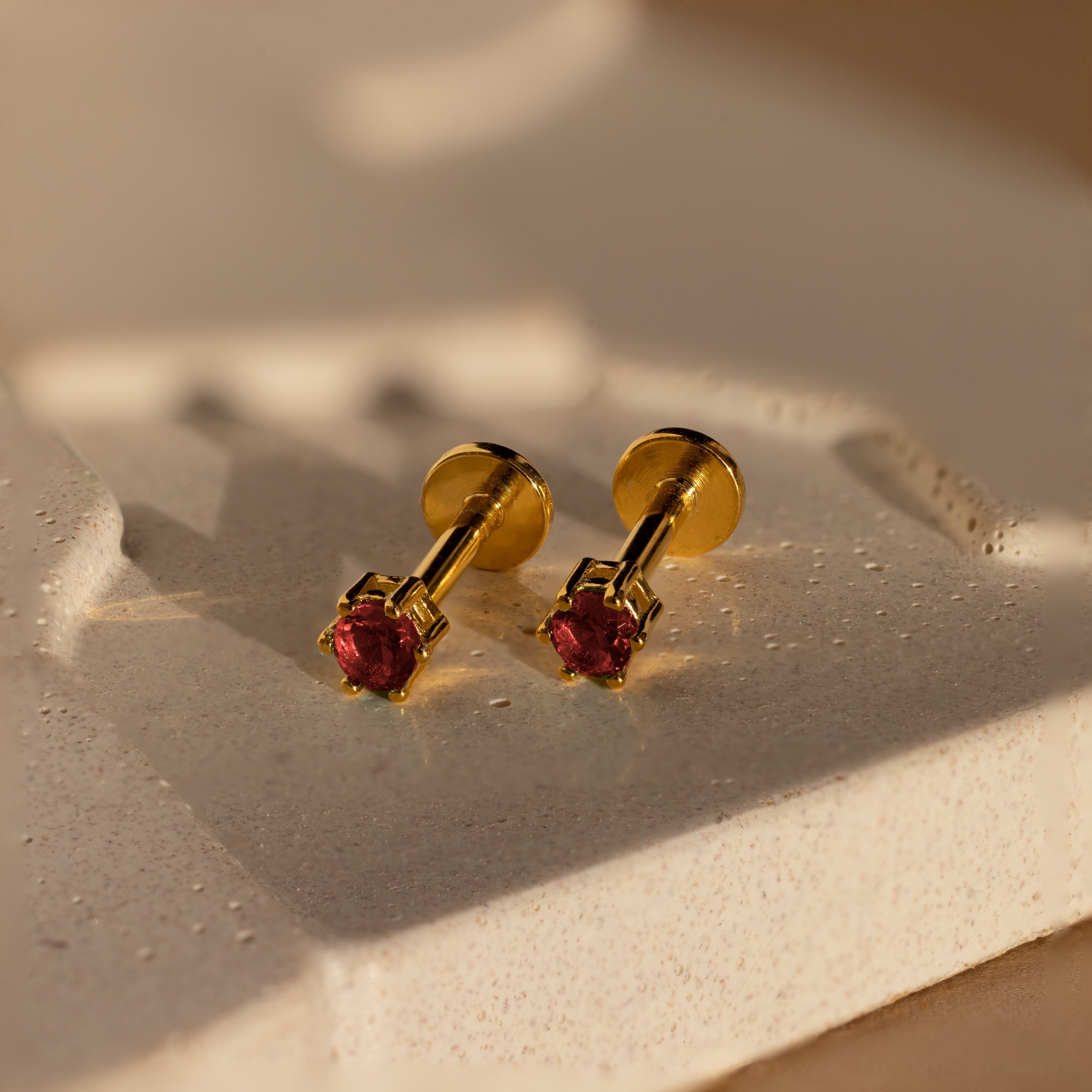 Elegant Garnet Flatback Studs with red gemstones, these gold earrings add a dainty touch and look stunning against a textured, light-colored surface.