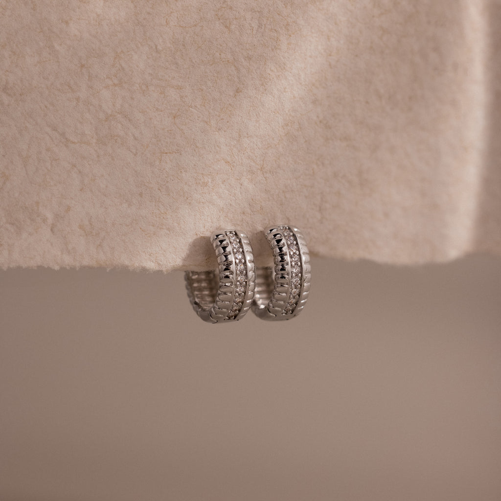 Ribbed Pave Huggies in Sterling Silver, featuring a textured, chunky hoop design, are displayed on a soft cream-colored fabric background.