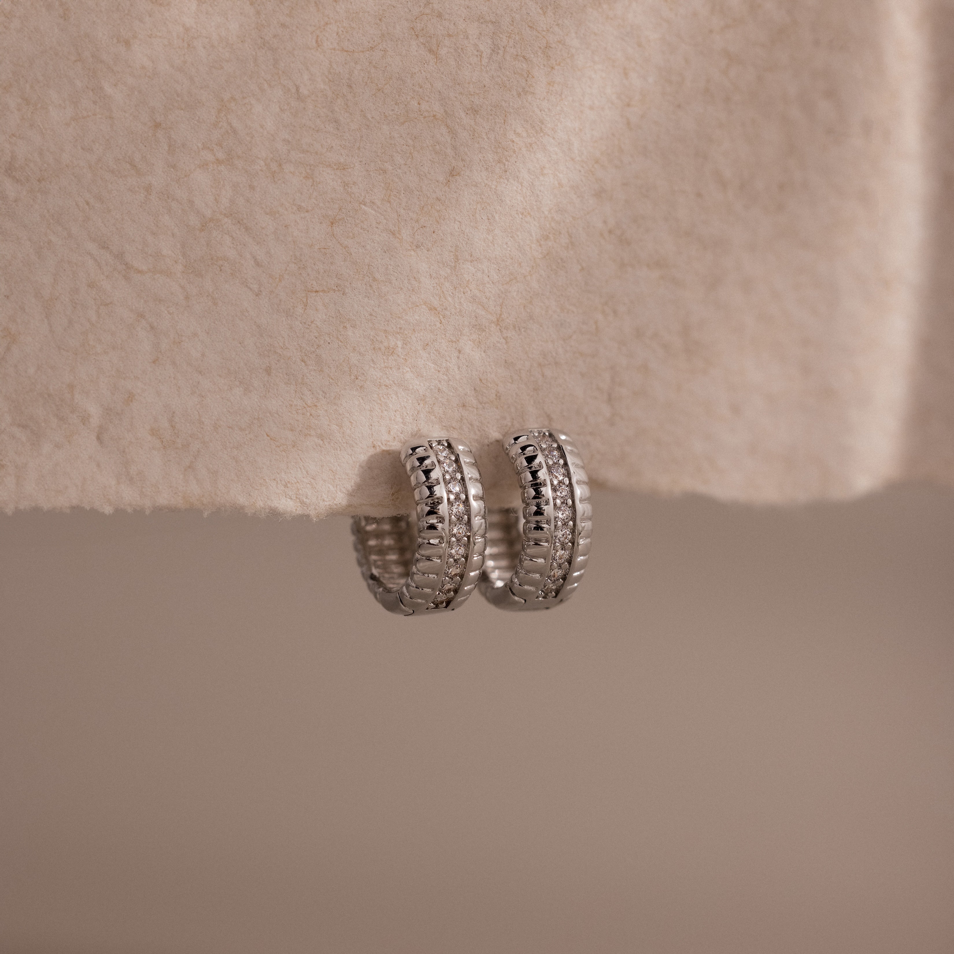 Ribbed Pave Huggies in Sterling Silver, featuring a textured, chunky hoop design, are displayed on a soft cream-colored fabric background.