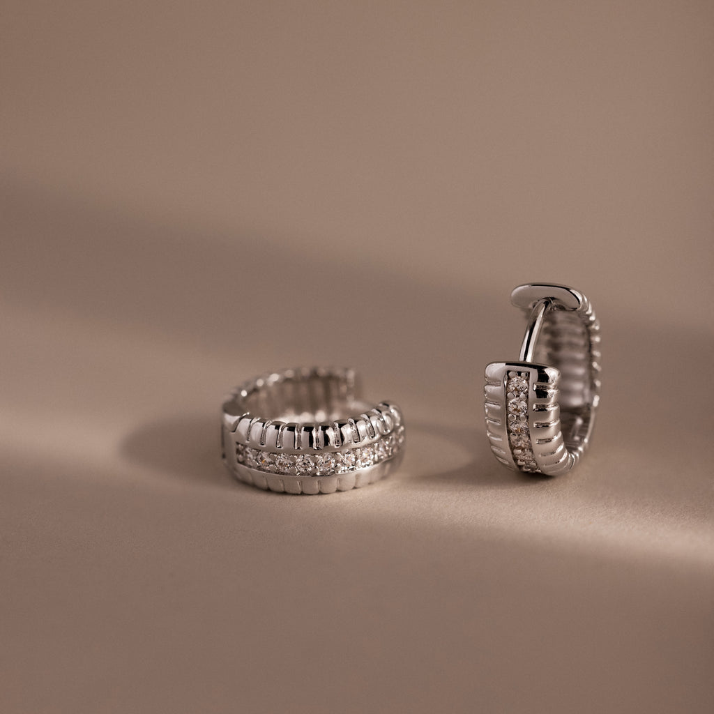 Two Ribbed Pave Huggies in Sterling Silver, featuring a row of sparkling stones, are displayed on a beige background.