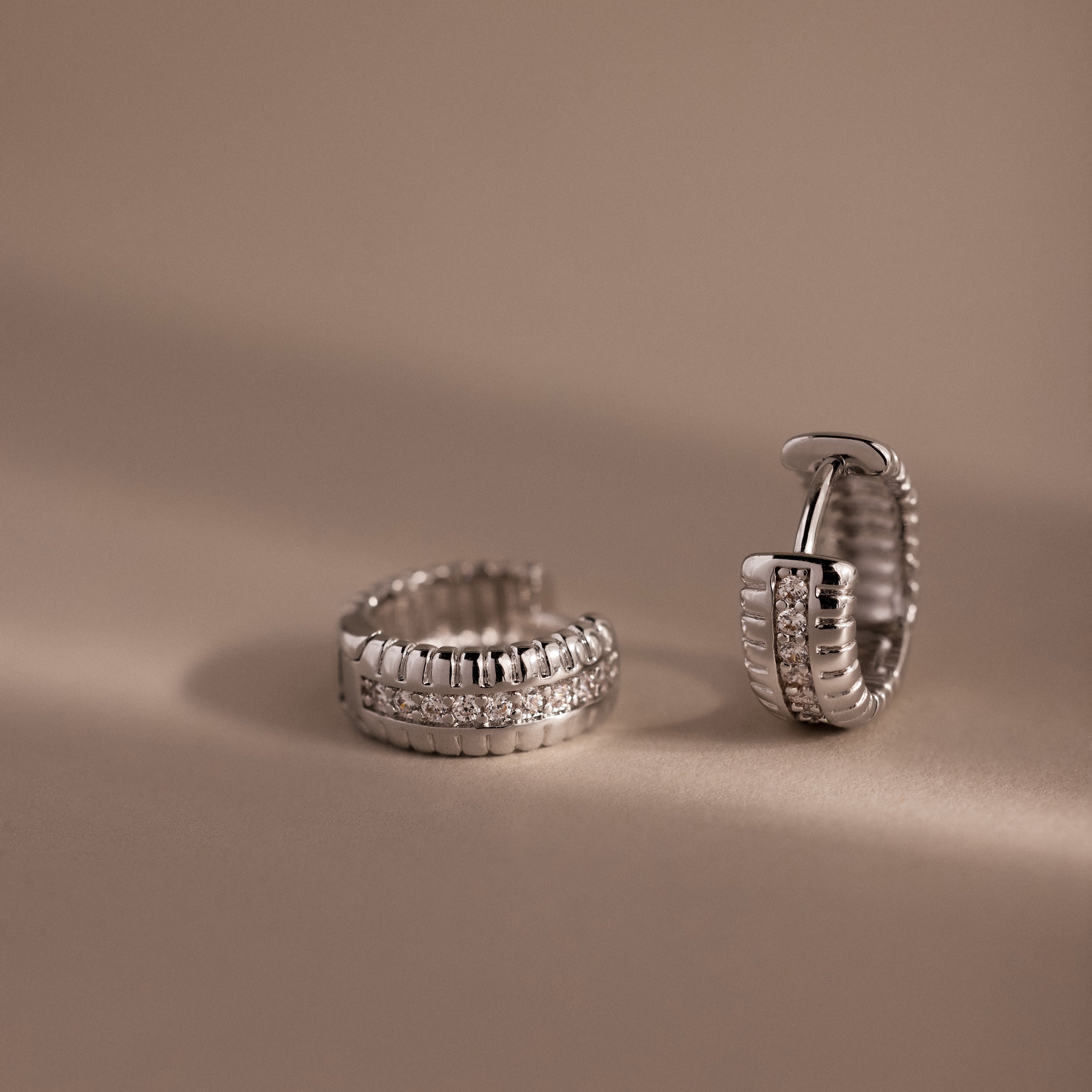 Two Ribbed Pave Huggies in Sterling Silver, featuring a row of sparkling stones, are displayed on a beige background.