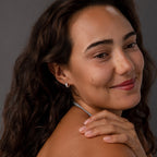 Smiling woman with wavy brown hair wears Ribbed Pave Huggies in Sterling Silver, touching her shoulder against a gray background.