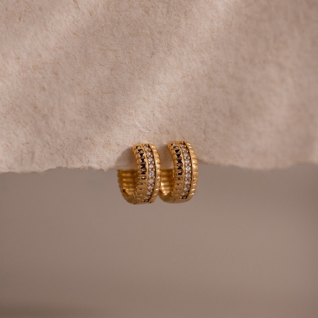 Ribbed Pave Huggies featuring a textured gold finish and embedded cubic zirconia stones, shown on a soft beige surface—ideal earrings for those who love statement jewelry.