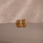 Ribbed Pave Huggies featuring a textured gold finish and embedded cubic zirconia stones, shown on a soft beige surface—ideal earrings for those who love statement jewelry.