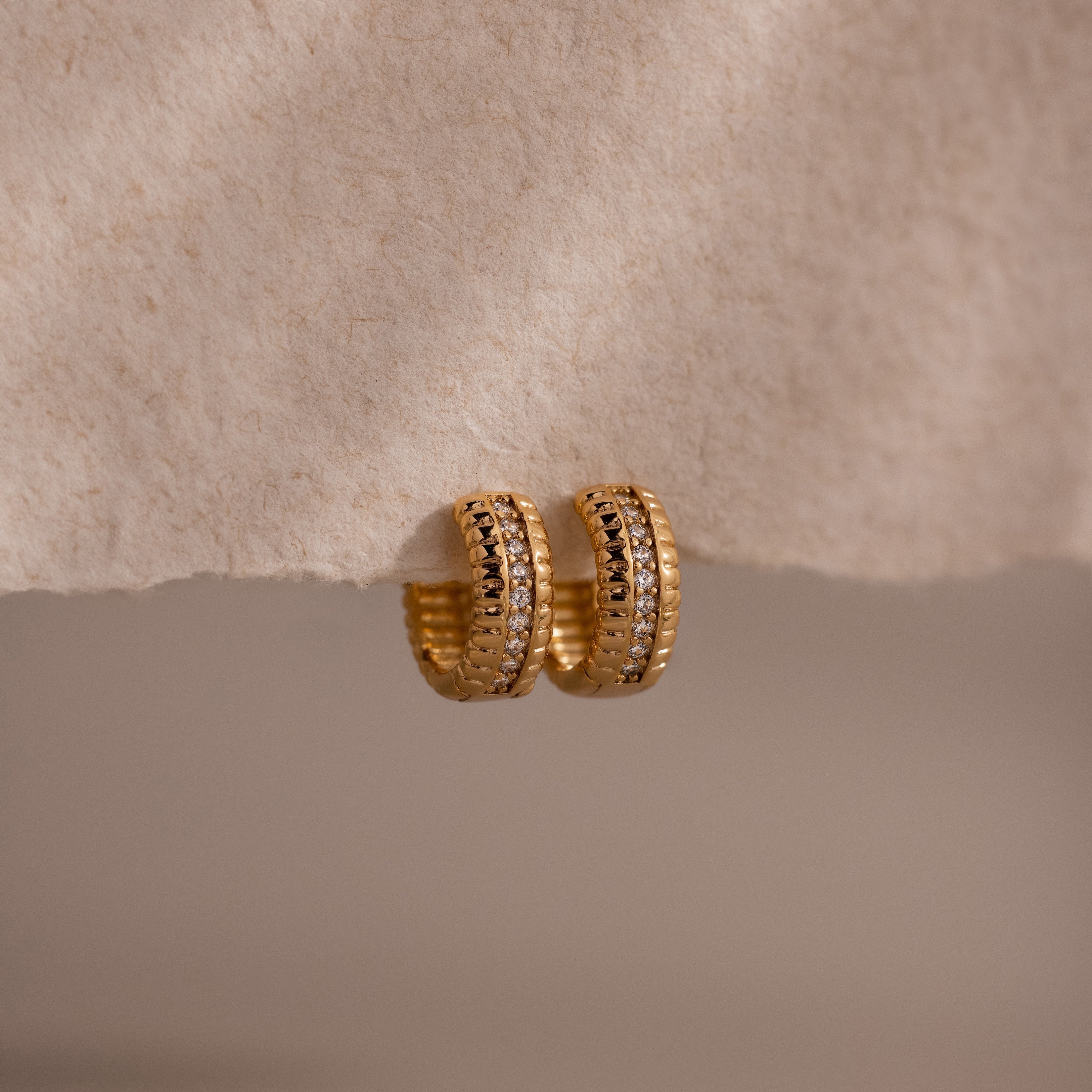 Ribbed Pave Huggies featuring a textured gold finish and embedded cubic zirconia stones, shown on a soft beige surface—ideal earrings for those who love statement jewelry.