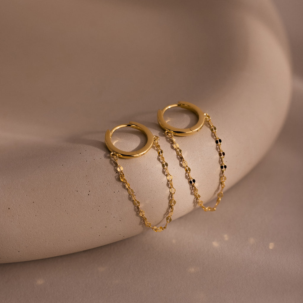 The Mirror Chain Huggies in 18K Gold feature gold hoops with delicate double chain details, perfect for minimalist jewelry lovers and shown on a smooth beige surface.