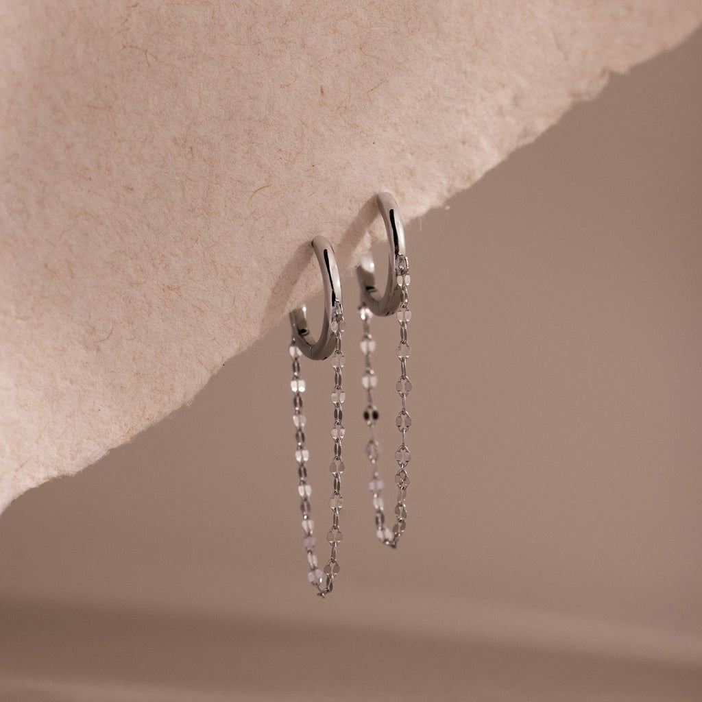 The Mirror Chain Huggies feature silver hoops with delicate chain drops, offering a minimalist jewelry style showcased elegantly on a textured beige surface.