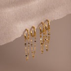 The Mirror Chain Huggies in 18K Gold, featuring delicate dangling chains, are displayed on a soft beige textured surface—ideal for statement earring lovers and fans of minimalist jewelry.