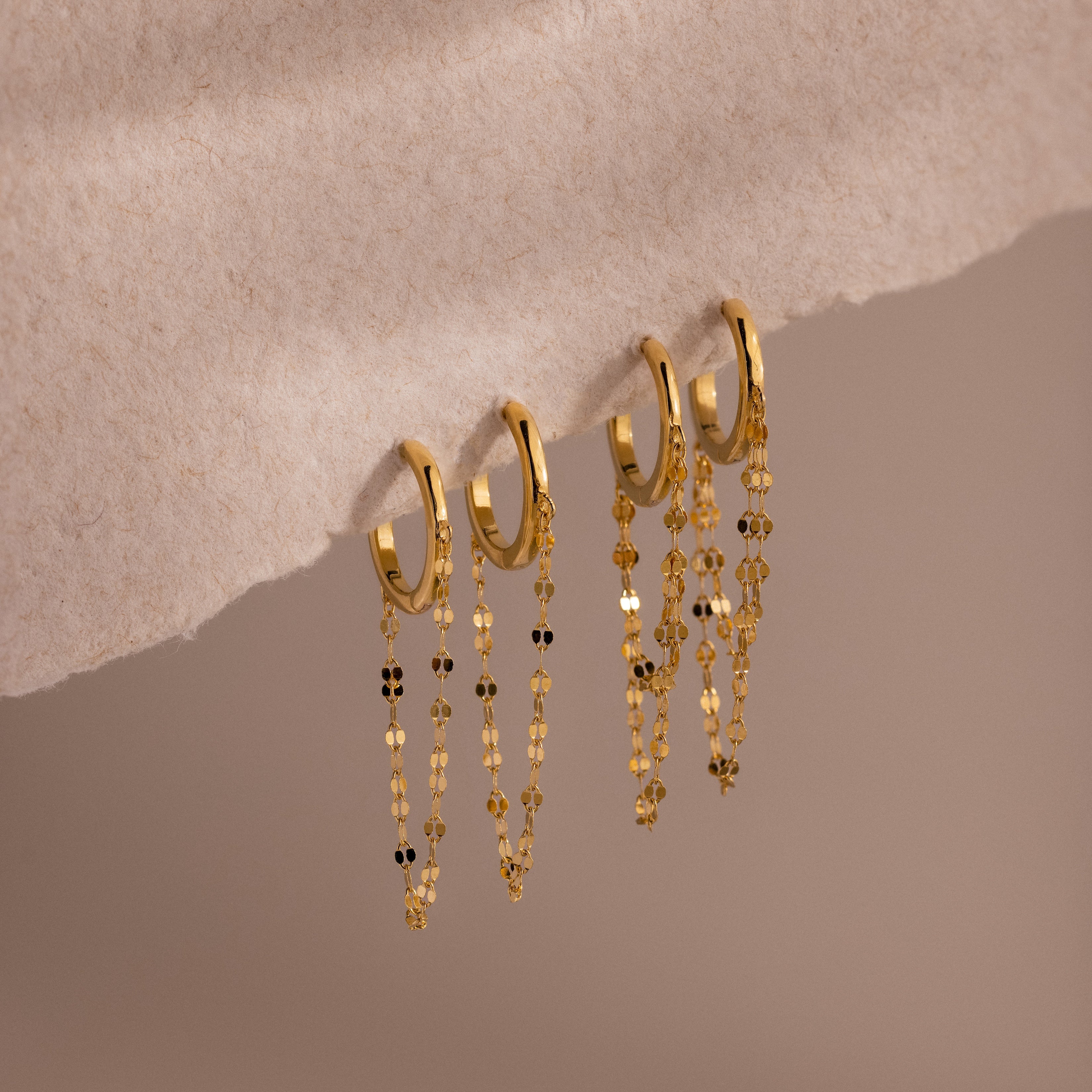 The Mirror Chain Huggies in 18K Gold, featuring delicate dangling chains, are displayed on a soft beige textured surface—ideal for statement earring lovers and fans of minimalist jewelry.