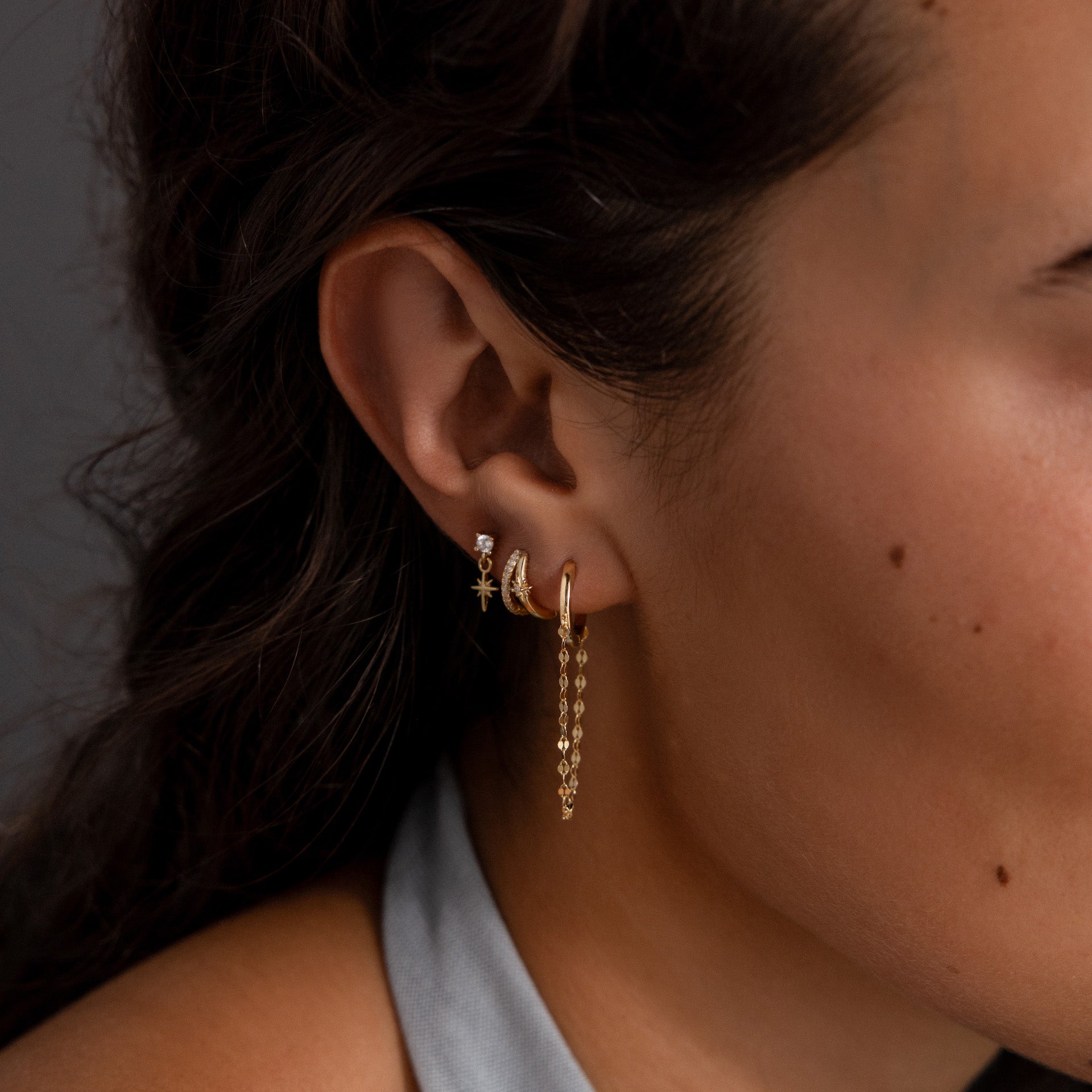 Close-up of a woman's ear with wavy dark hair, styled with three gold hoops and a chain earring. The look is elevated by the Starburst Pave Duo Huggies in 18K Gold, adding celestial sparkle and modern elegance.