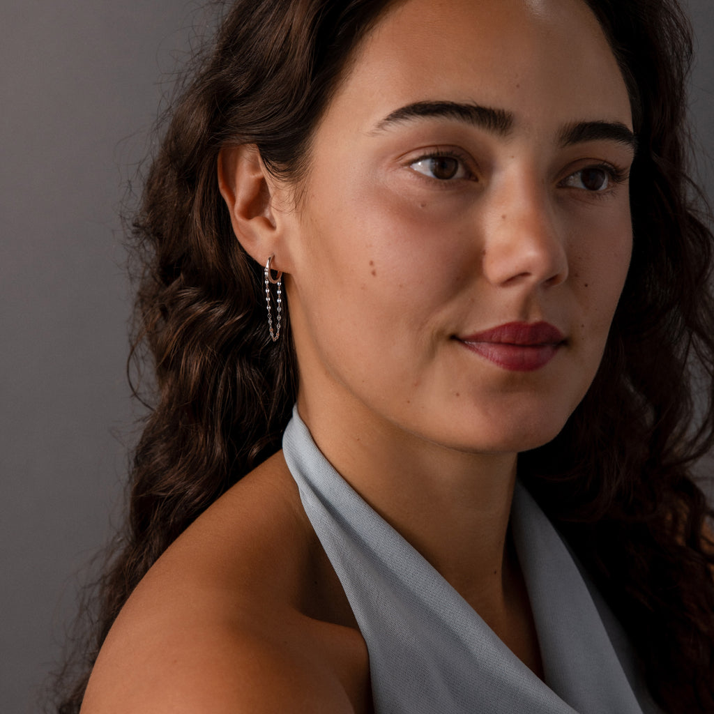 Woman with long curly hair wearing a grey halter top and Mirror Chain Huggies in Sterling Silver, looking off to the side.