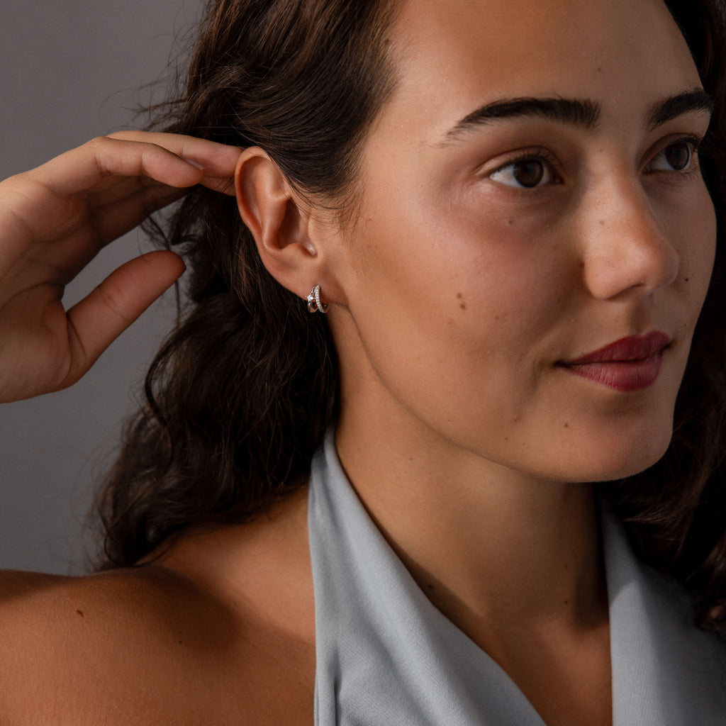 Woman with wavy brown hair and silver earrings, wearing a light gray halter top, touches her ear adorned with Pave Diamond Duo Huggies in Sterling Silver.