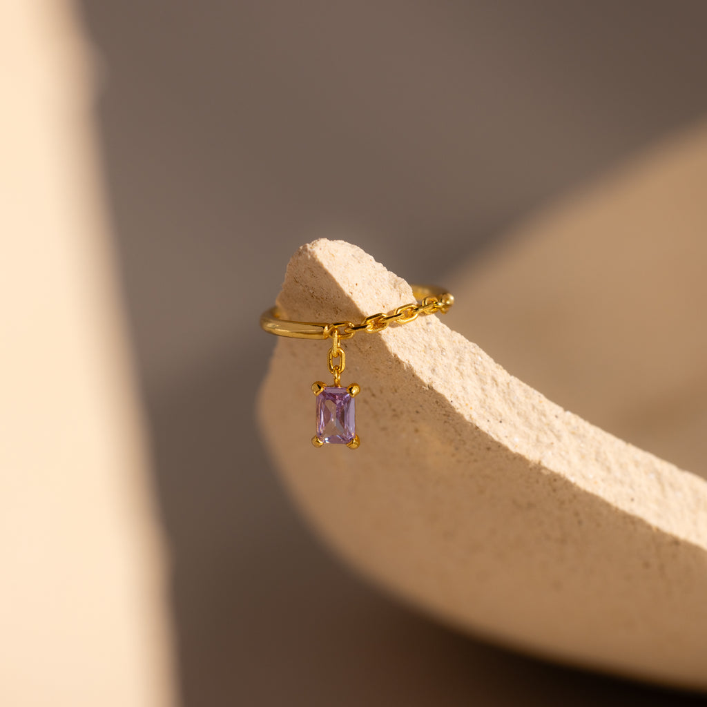 Alexandrite Drop Chain Ring featuring a dangling, emerald-cut purple gem rests on the edge of a textured beige ceramic surface.