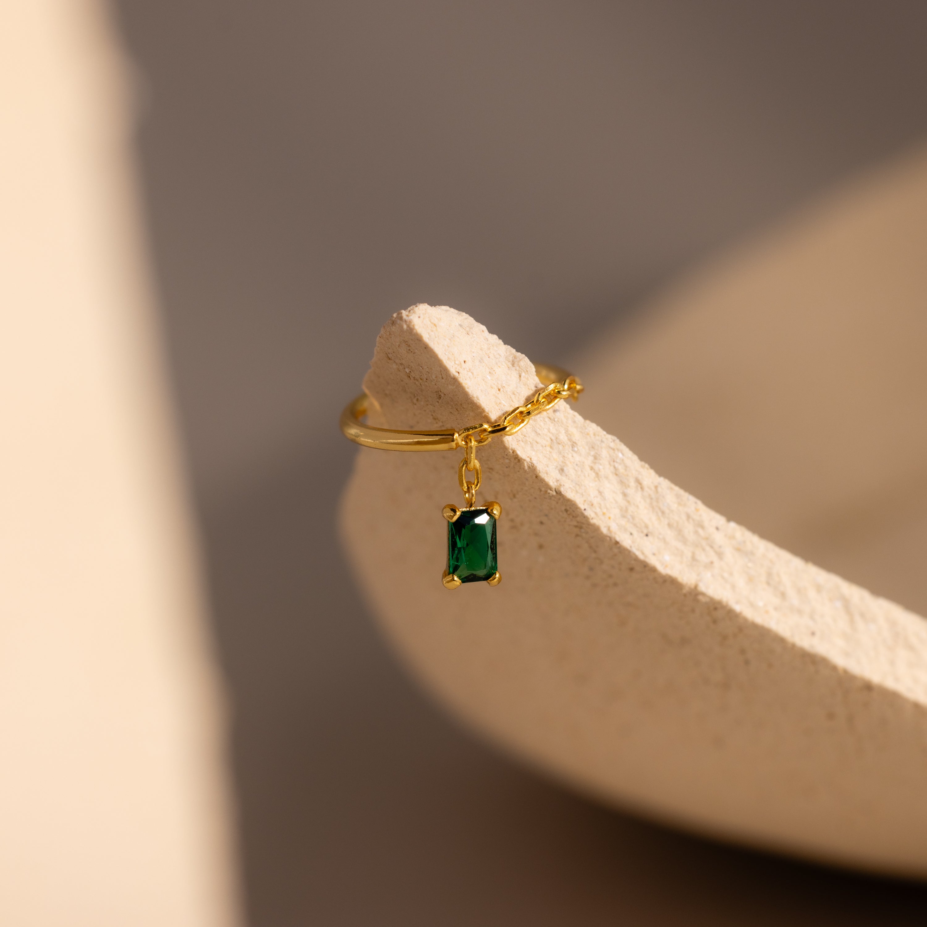 The Emerald Drop Chain Ring features a delicate gold chain with a small emerald-cut green gemstone charm, elegantly displayed on a textured beige ceramic piece.