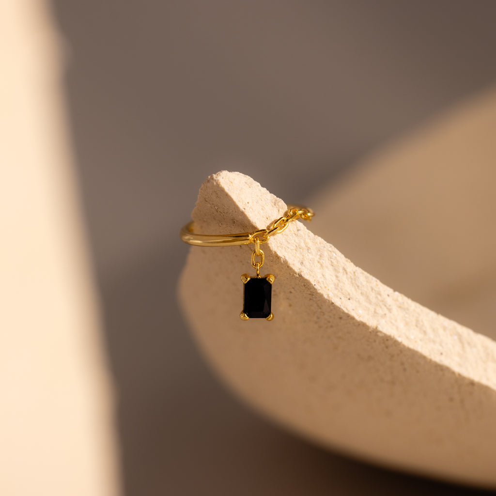 Onyx Drop Chain Ring featuring a small emerald-cut black stone charm, displayed on a textured beige surface.