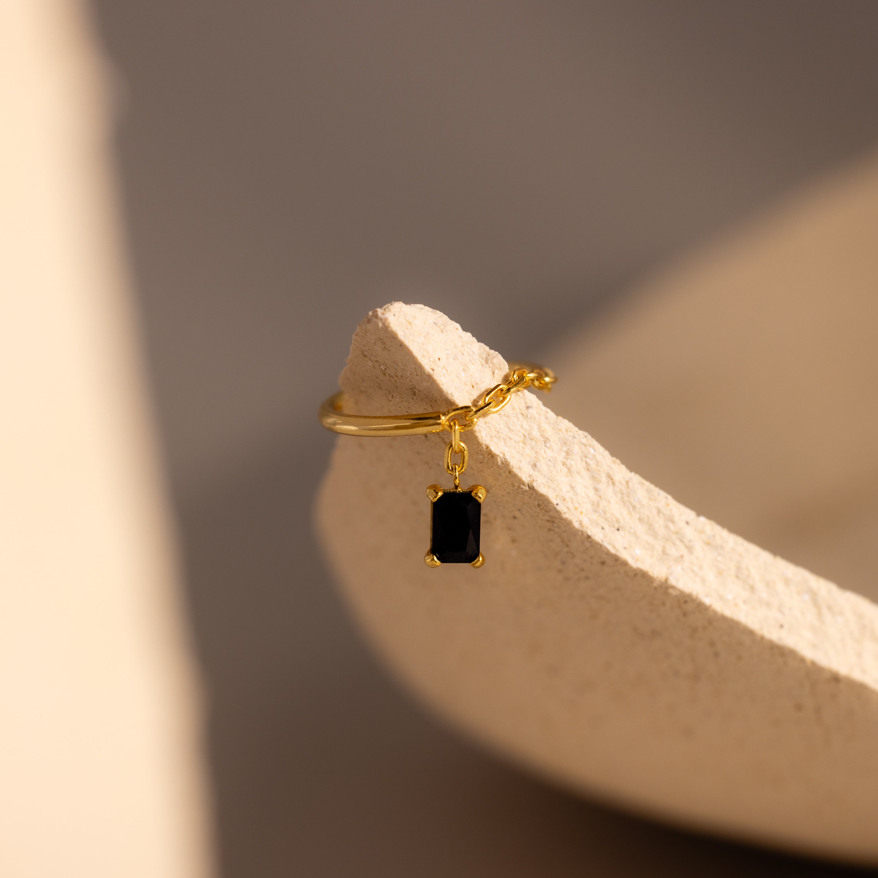 Onyx Drop Chain Ring featuring a small emerald-cut black stone charm, displayed on a textured beige surface.