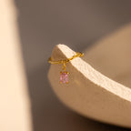The Pink Tourmaline Drop Chain Ring features a delicate gold band with a pink tourmaline pendant, elegantly displayed on the edge of a textured beige surface.