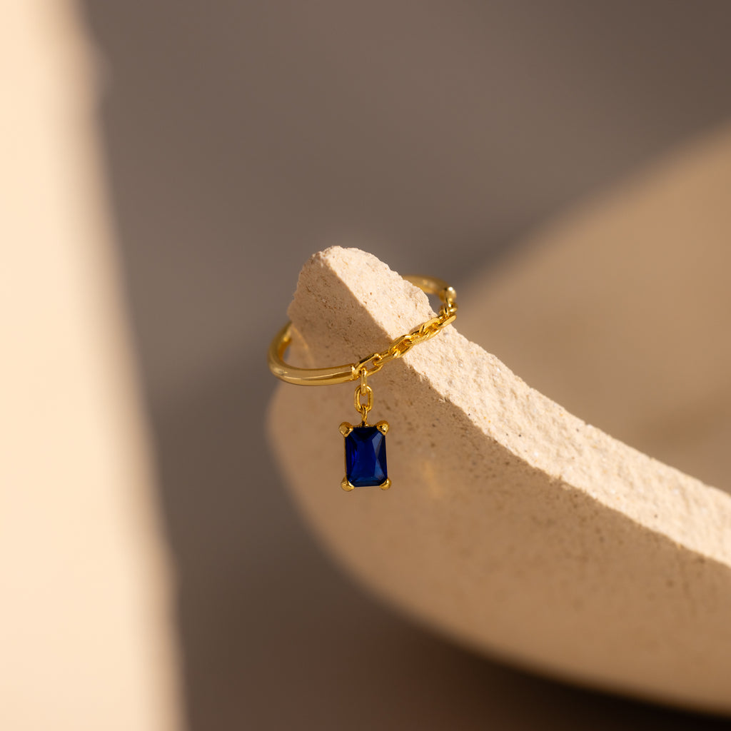 Sapphire Drop Chain Ring with an emerald-cut blue gemstone charm, shown on a textured beige surface in soft light.