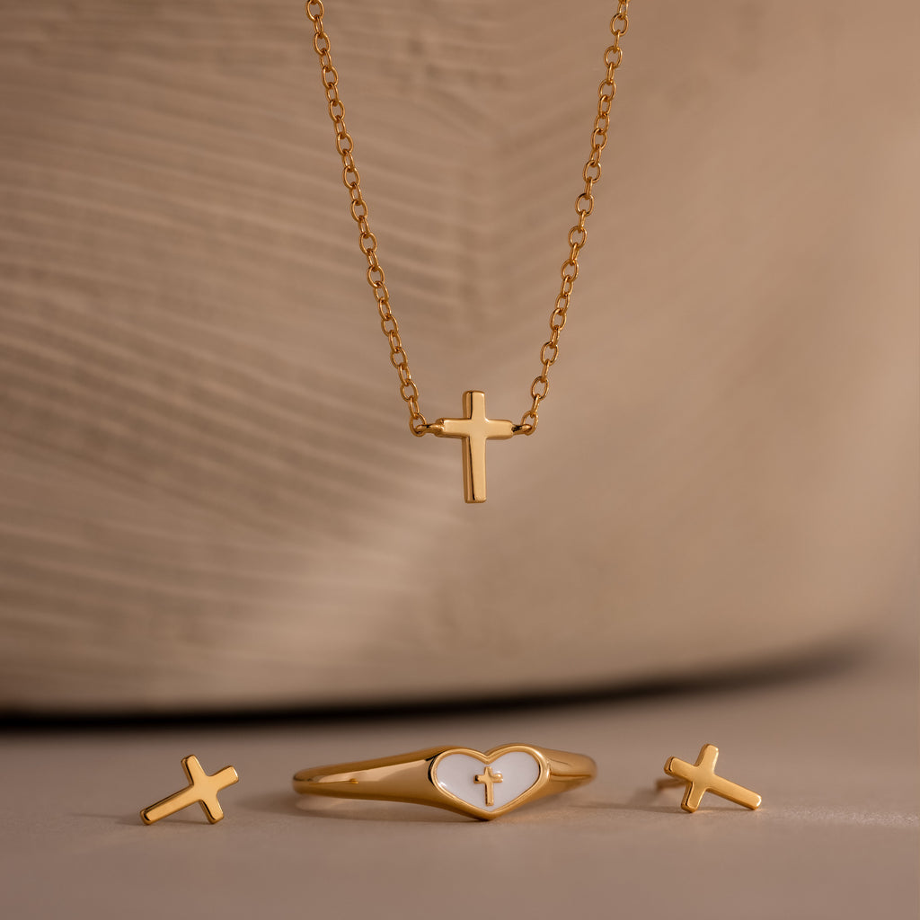 Gold cross jewelry set featuring a delicate cross pendant necklace, matching cross stud earrings, and a gold heart ring with white enamel and cross detail, displayed against a beige background.
