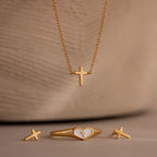 Gold cross jewelry set featuring a delicate cross pendant necklace, matching cross stud earrings, and a gold heart ring with white enamel and cross detail, displayed against a beige background.