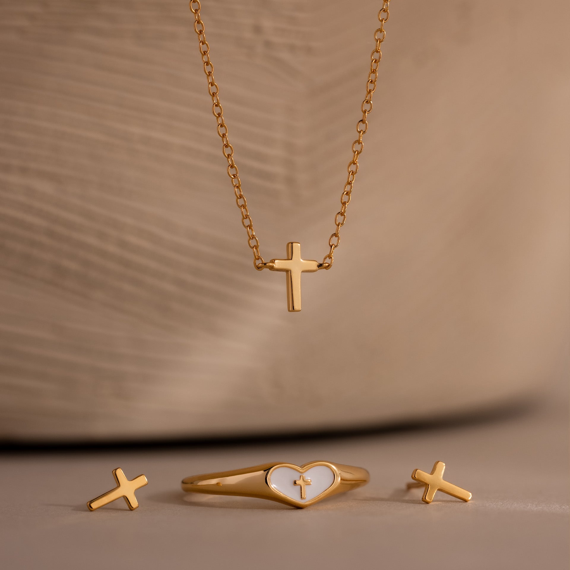Gold cross jewelry set featuring a delicate cross pendant necklace, matching cross stud earrings, and a gold heart ring with white enamel and cross detail, displayed against a beige background.