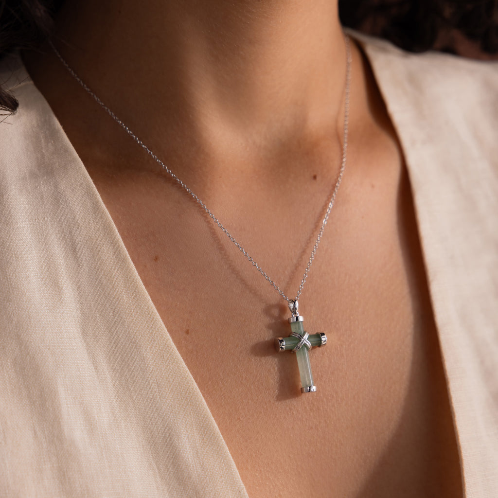 Silver chain necklace with a green jade cross pendant featuring polished gold end caps and a gold X shaped accent, worn with an ivory V neck blouse.