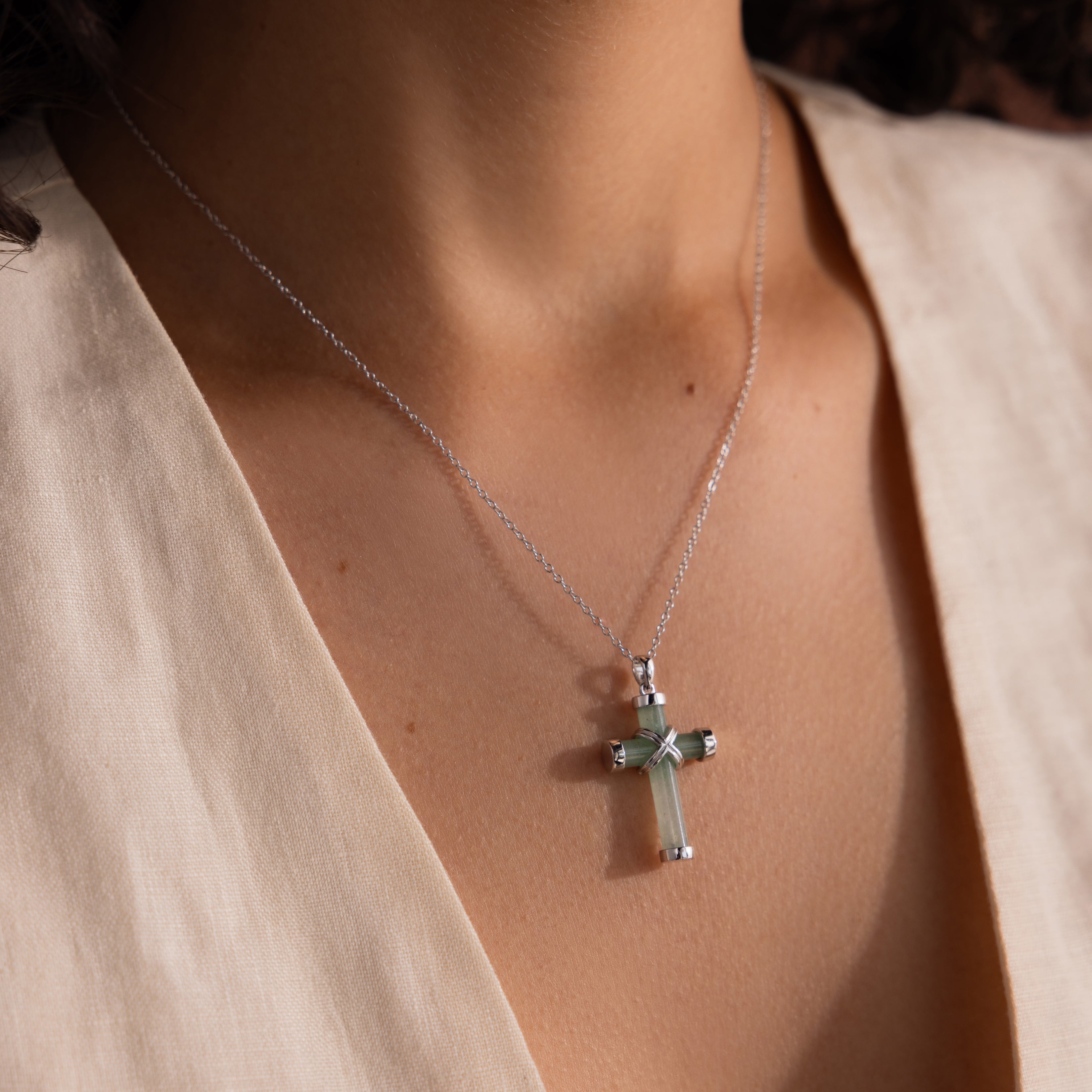 Silver chain necklace with a green jade cross pendant featuring polished gold end caps and a gold X shaped accent, worn with an ivory V neck blouse.