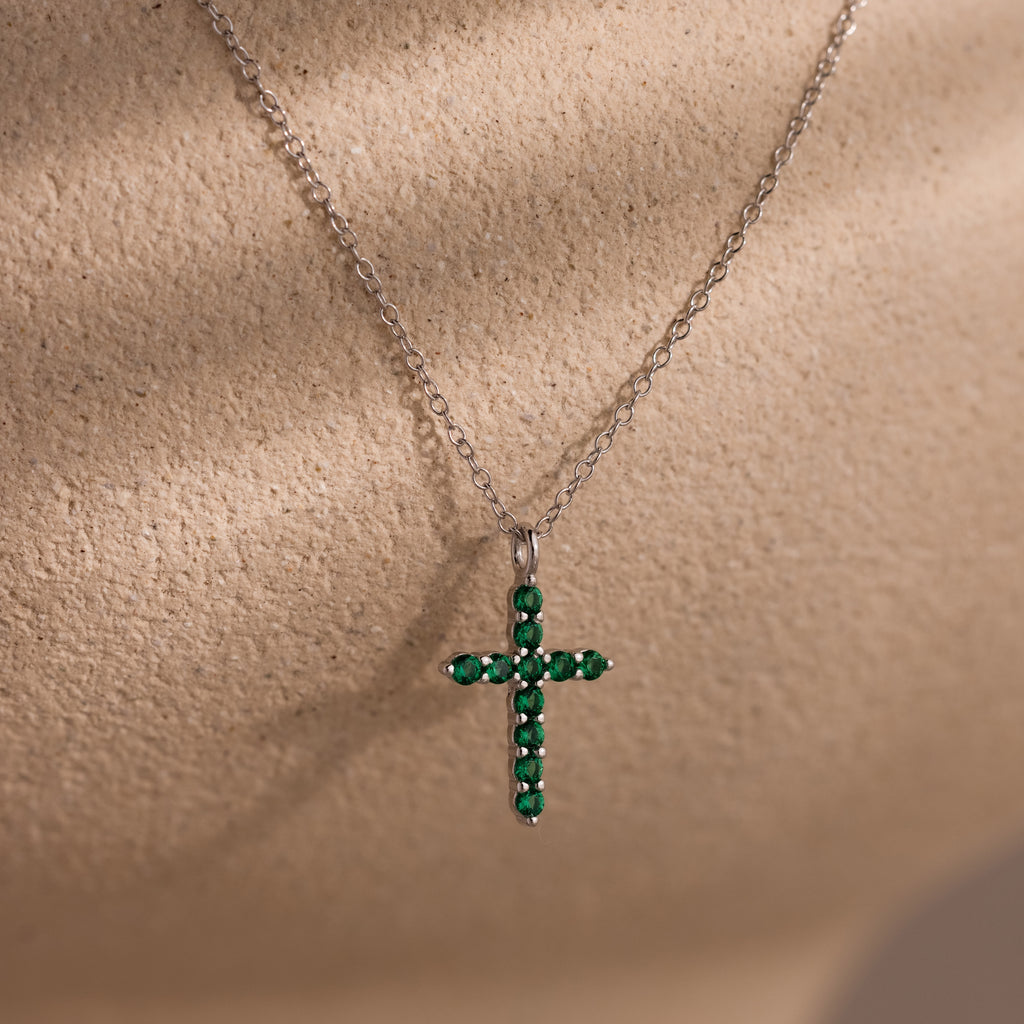 Silver chain necklace featuring a cross pendant adorned with small round green gemstones, displayed against a textured beige background.