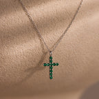 Silver chain necklace featuring a cross pendant adorned with small round green gemstones, displayed against a textured beige background.