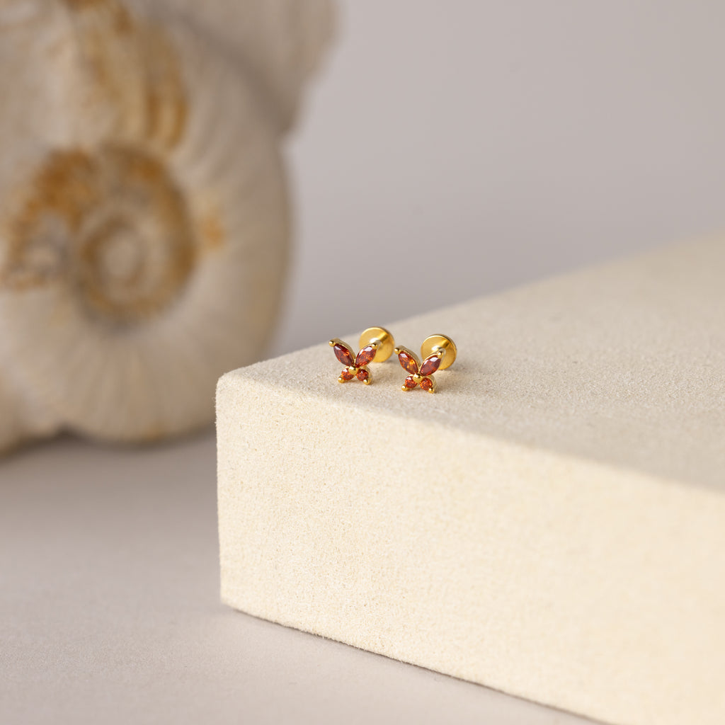 Butterfly Garnet Flatback Studs—small gold earrings with pink gemstones—are displayed on a beige surface, accented by a seashell in the background.