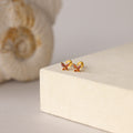 Butterfly Garnet Flatback Studs—small gold earrings with pink gemstones—are displayed on a beige surface, accented by a seashell in the background.