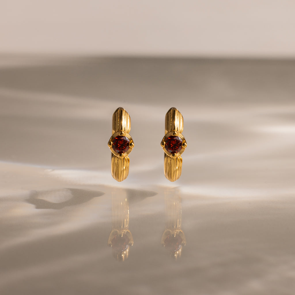 The Ribbed Garnet Huggies, gold hoop earrings with red gemstones, stand upright on a softly lit reflective surface—making them a perfect meaningful gift.